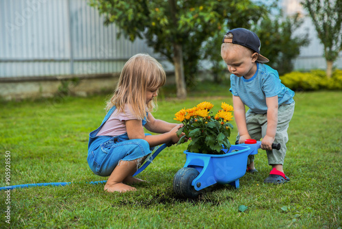 Cute little boy holding wheelbarrow while his sister watering flowers outdoors