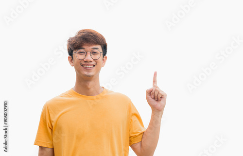 A young Asian man in his 20s wearing glasses and wearing a yellow t-shirt happy smiling pointing thumbs up isolated on a gray background. The person's index finger pointing to a blank space for text