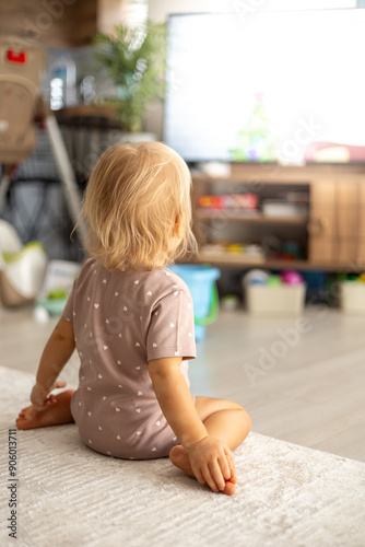 A small blonde baby girl sitting on the floor at home and wathing TV photo