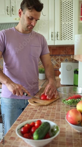 Young handsome happy man in casual wear is preparing wonderful fresh vegan salad from ripe summer vegetables in the kitchen at home	