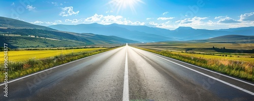 A straight empty road stretches into the horizon under a bright sunny sky, surrounded by green fields and distant mountains.
