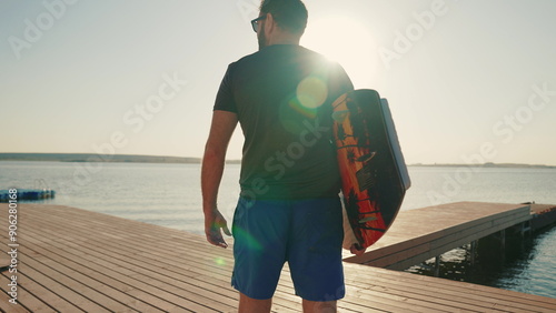 A man with a wakeboard walking on a lake pier on a sunny summer day. Male surfer holding a short surfboard while walking on the pier.