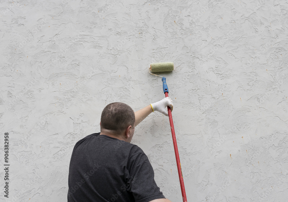 A man diligently applies fresh paint to a textured wall, showcasing skill and determination under the open sky. A man diligently applies fresh paint to a textured wall, showcasing skill and determination under the open sky.