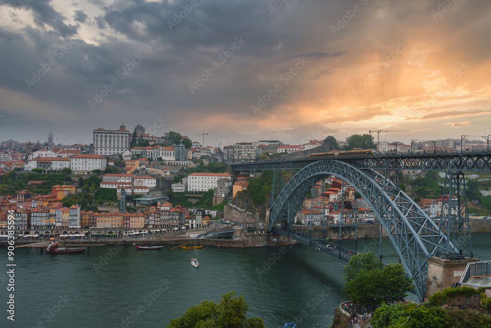 Naklejka premium Iconic Dom Luis I Bridge spans the Douro River in Porto, Portugal, connecting the city's banks. Ribeira district's colorful buildings and dramatic sky enhance the scene.
