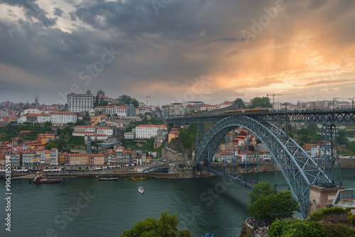 Iconic Dom Luis I Bridge spans the Douro River in Porto, Portugal, connecting the city's banks. Ribeira district's colorful buildings and dramatic sky enhance the scene.