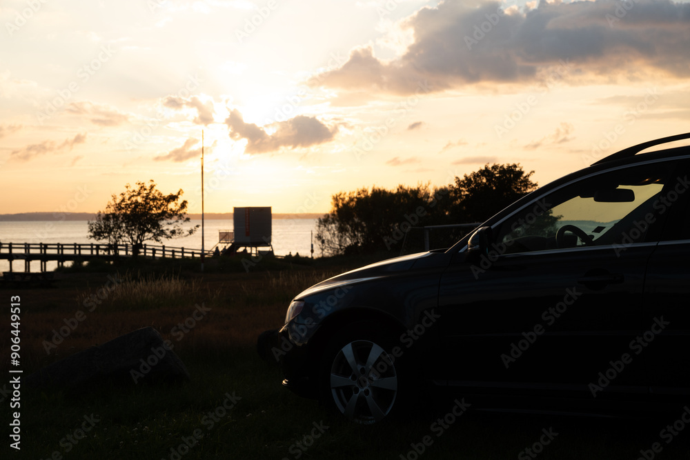 Fototapeta premium Car camping in Skåne south of Helsingborg. A beautiful midsummer nights sunset at the beach. Light mirroring in freshly polished car. dramatic clouds.