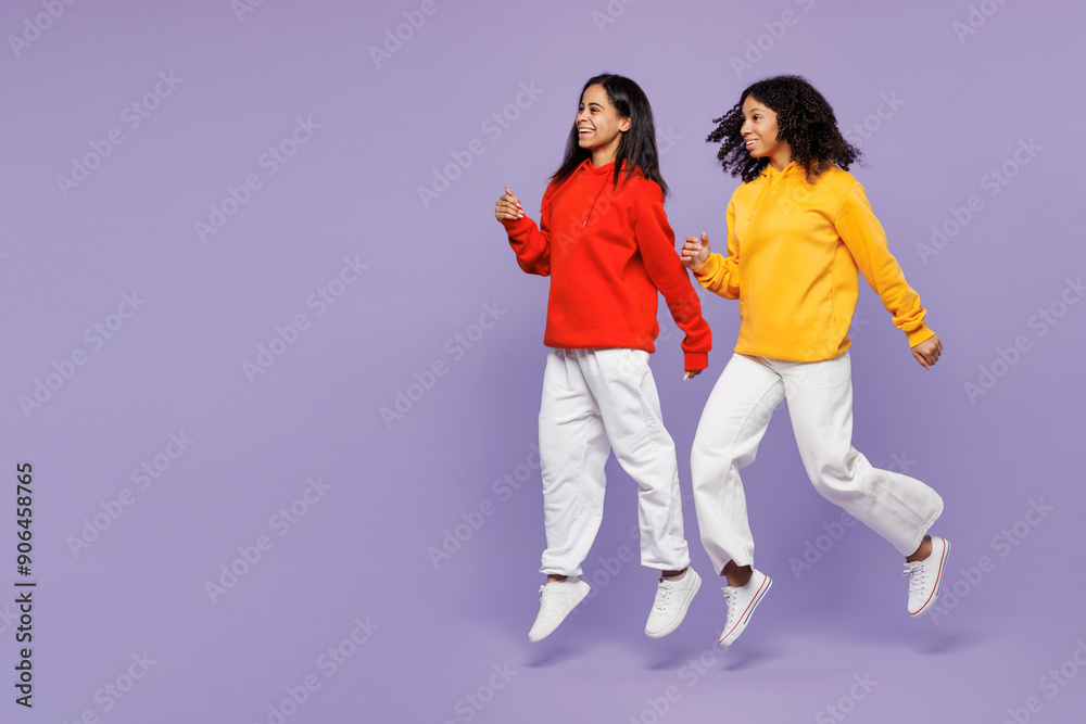 Obraz premium Full body side view two young happy little kid teen girls women they wear red yellow hoodie casual clothes jump high run fast look aside isolated on plain pastel purple background. Lifestyle concept.