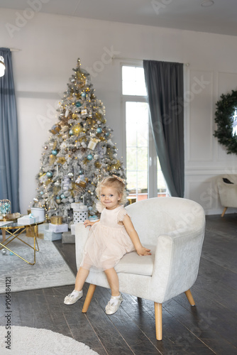 smiling positive young girl toddler 2 years old wearing pink dress sits white armchair decorated Christmas tree. tree decorated with gold, silver, and blue ornaments. concept: celebrating new year 