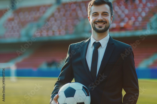 A confident man in a suit and tie stands on a soccer field, holding a soccer ball, with empty stadium seats in the background. photo