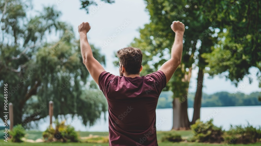 A man stands with arms raised in celebration, overlooking a tranquil lake surrounded by trees on a sunny day, capturing a moment of victory and joy