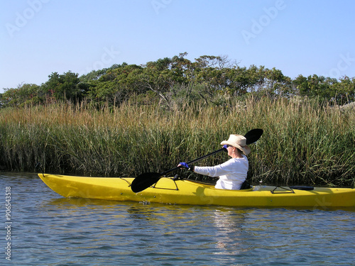 A middle aged woman is paddling a yellow kayak along a river with marshes in the background