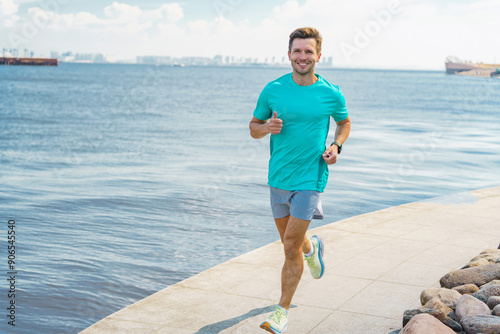 Smiling man jogging along a waterfront path, wearing a turquoise shirt and shorts, exuding energy and fitness.