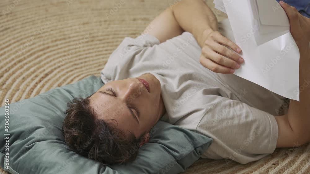 Focused adult man college student laying on the floor in apartment room at daylight and doing homework, stressed throws paper sheets in the air. Education concept. Focused adult man college student laying on the floor in apartment room at daylight and doing homework, stressed throws paper sheets in the air. Education concept.