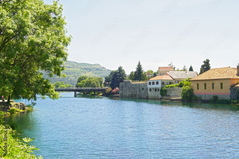 Fototapeta premium Summer landscape from the center of Trebinje: the Trebišnjica River, trees, houses of the Old Town on the bank and a mountain in the distance. Tourist places in Bosnia and Herzegovina.