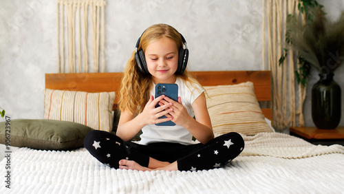 Little girl enjoying her phone and headphones while relaxing on the bed at home. A charming scene that captures the joy of childhood and modern technology, ideal for showcasing family life and enterta photo