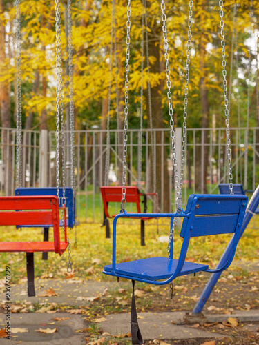 Empty old-fashioned merry-go-round in a city park. Red and blue wooden seats on metal chains of swing carousel. Trees on the background, fallen leaves on the ground.