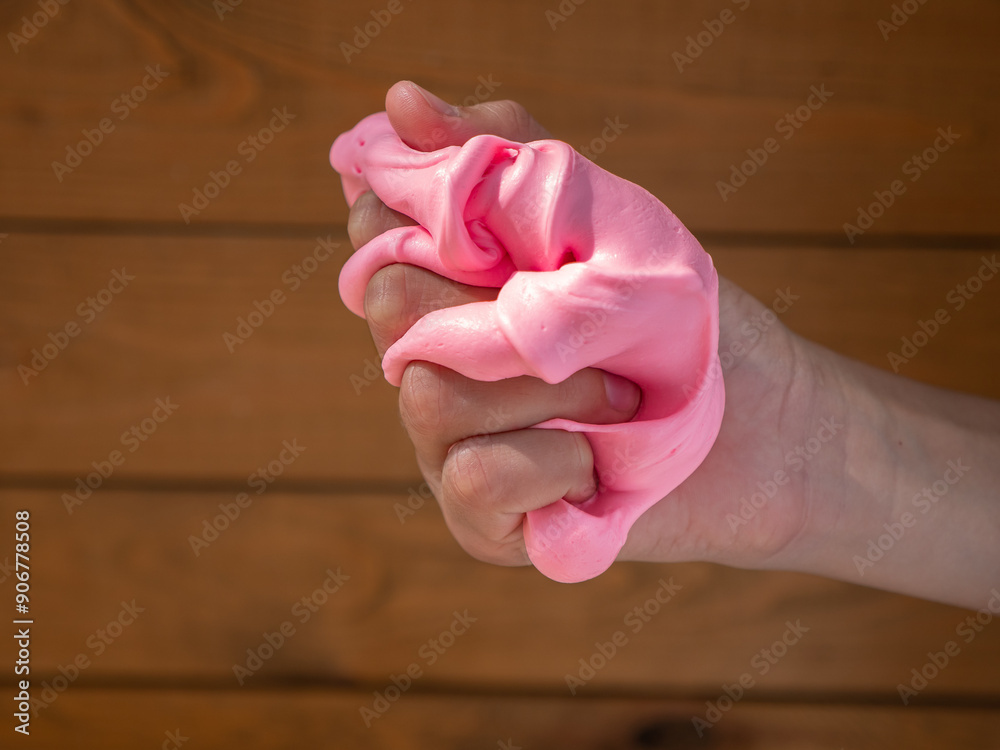 Child hand holding and squeezing fluffy soft pink slime against brown wooden background.