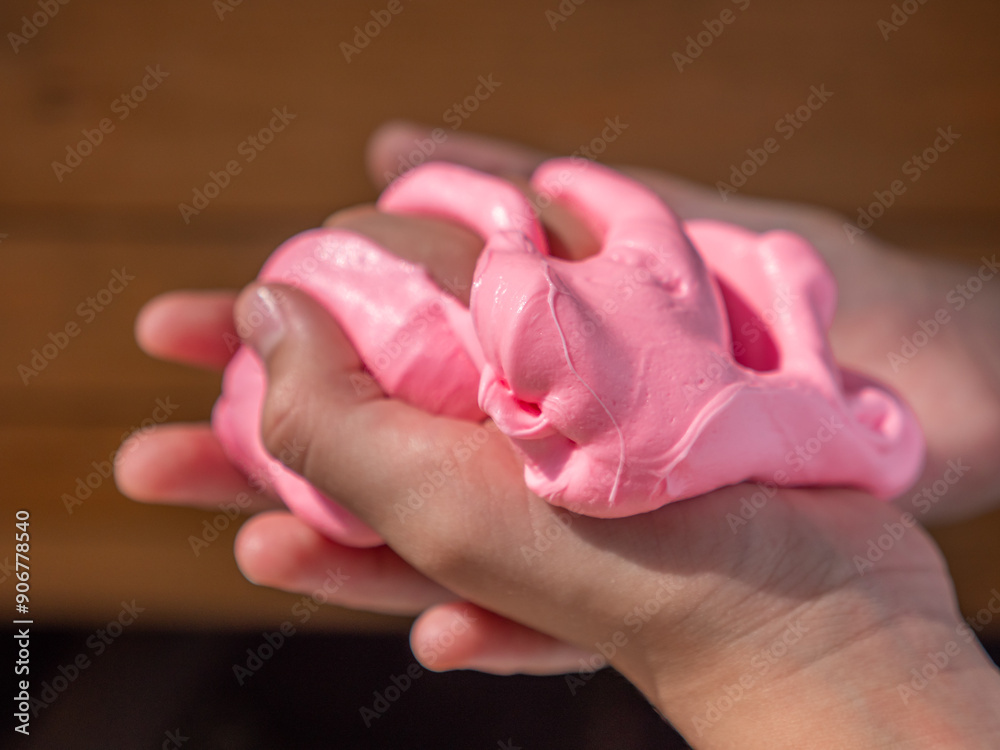 Child hand holding and squeezing fluffy soft pink slime against brown wooden background.