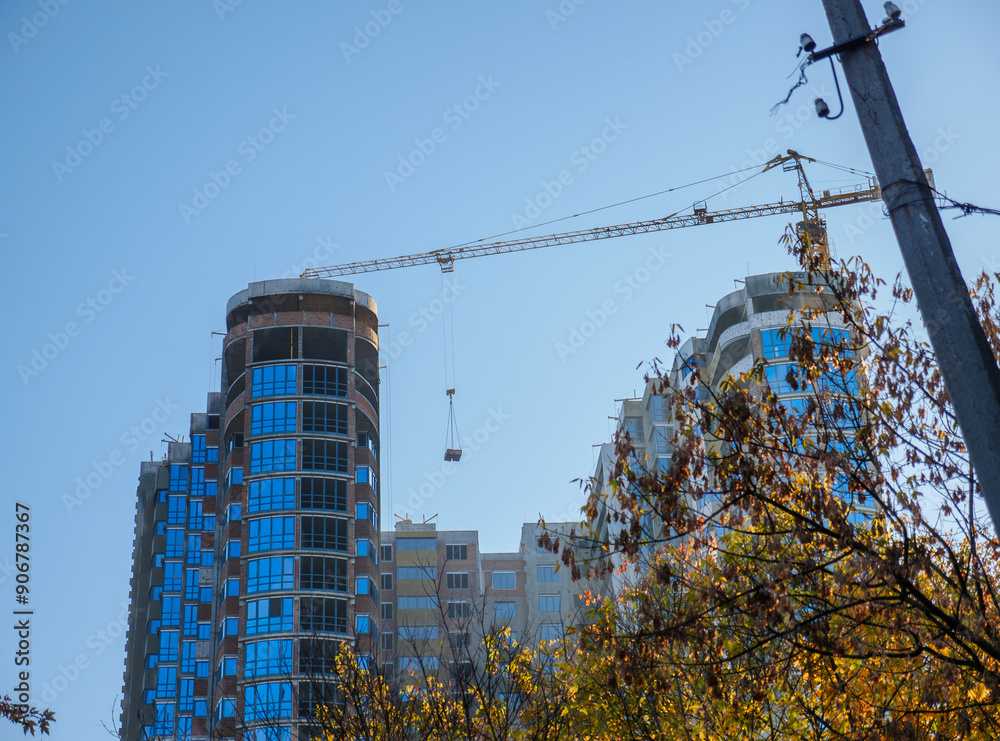 A crane lifts a load to the new buildings at the construction site, a tree and an electric pole in the foreground. New district under construction in Kyiv, Ukraine.