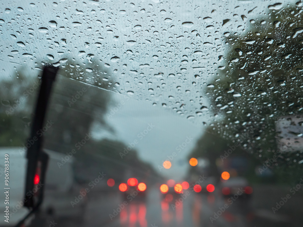 Closeup of raindrops on a windshield, blurry wipers cleaning the glass, road traffic with blurry car lights. View from the driver's seat.