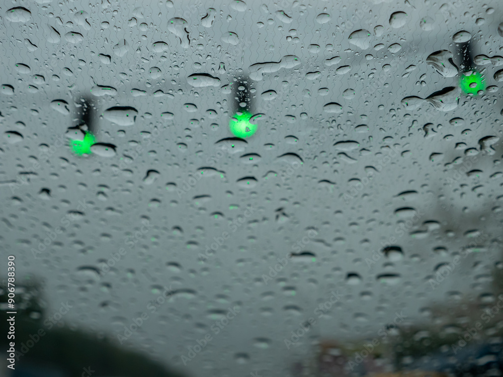 Closeup of rain drops on a windshield, blurry traffic lights with green signals directing traffic. View from the driver's seat.
