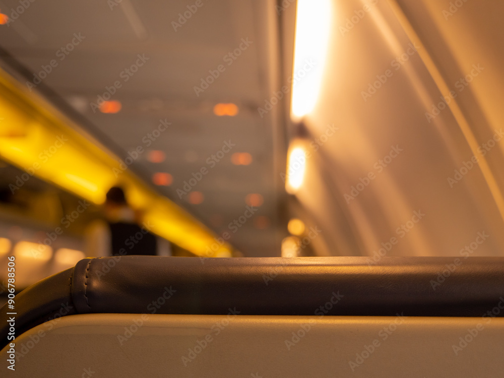 Blurry image of a steward standing in the aisle of an airplane aircraft cabin. View from behind the seat. Yellow lights turned on.