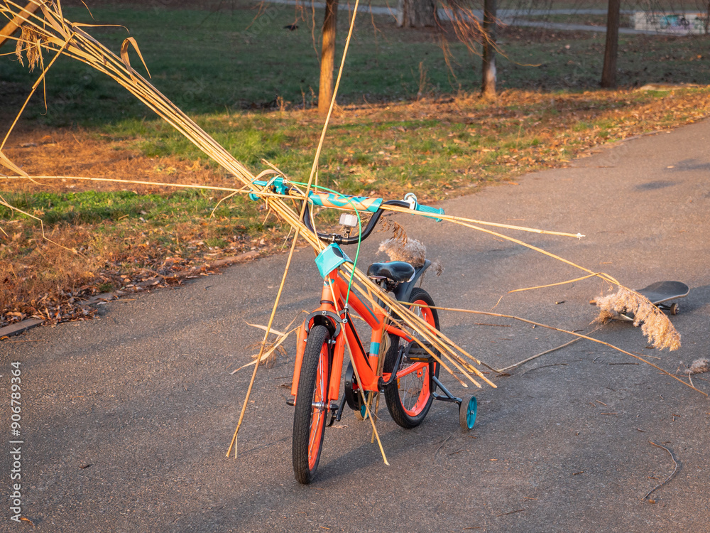 Two-wheeled orange and blue child bike with extra wheels carrying bulrush and pulling a skate board on a rope behind. Child playing active creative games outdoors in a park.