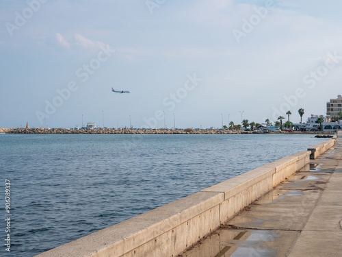 Obraz na płótnie Airplane preparing to land at the airport of Larnaca, Cyprus. Flying low above calm smooth blue mediterranean sea with rocky wave breaker and promenade. w salonie