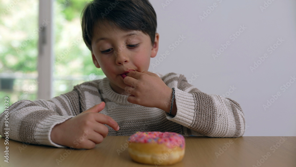 Young boy delaying gratification but tempted to eat a colorful sprinkled donut, grabbing sprinkles surrounding it with a focused look, creating a playful and delightful moment Young boy delaying gratification but tempted to eat a colorful sprinkled donut, grabbing sprinkles surrounding it with a focused look, creating a playful and delightful moment