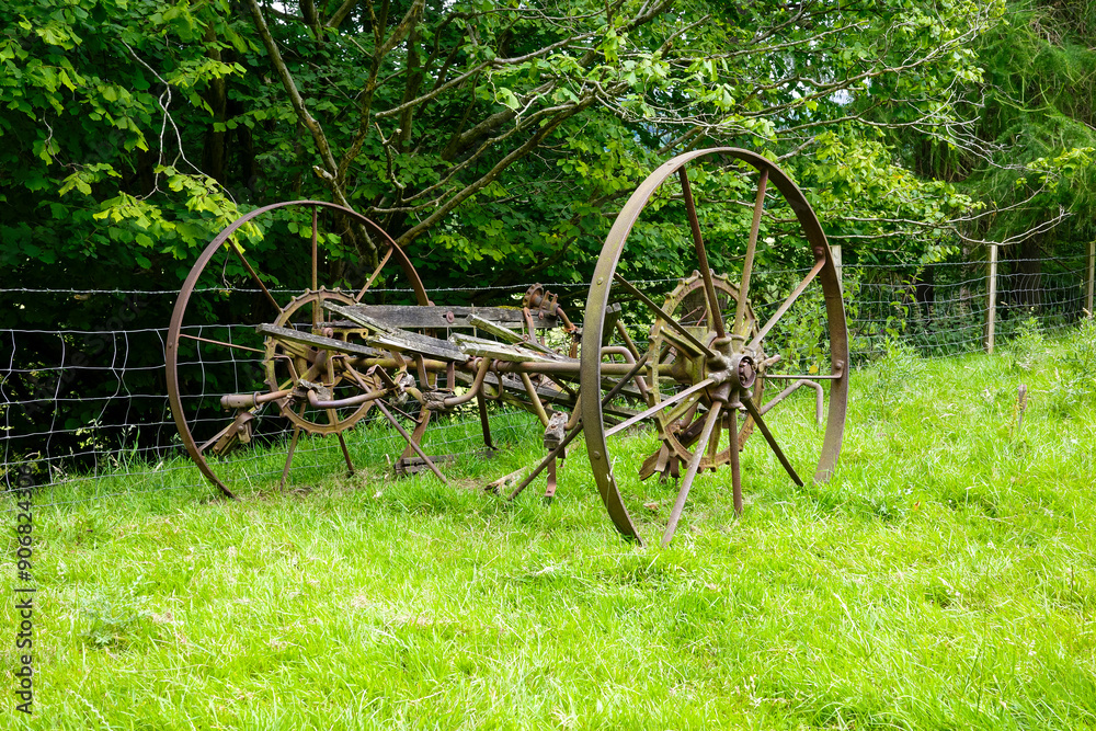 Rustic old farming equipment in a lush green field surrounded by trees and a wire fence. Rustic old farming equipment in a lush green field surrounded by trees and a wire fence.