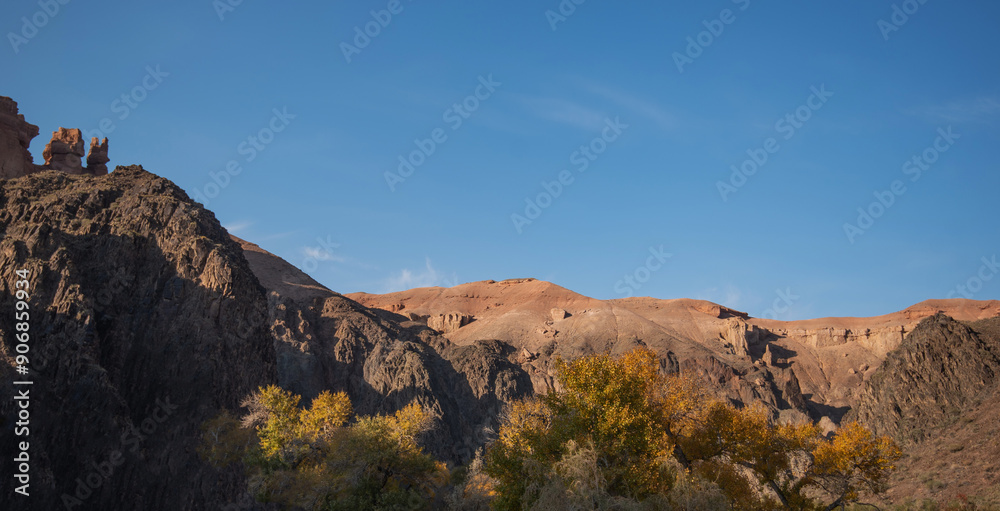 Naklejka premium autumn leaves contrast with the stark, imposing cliffs, as the soft glow of the setting sun bathes the serene desert landscape in a warm, golden light.