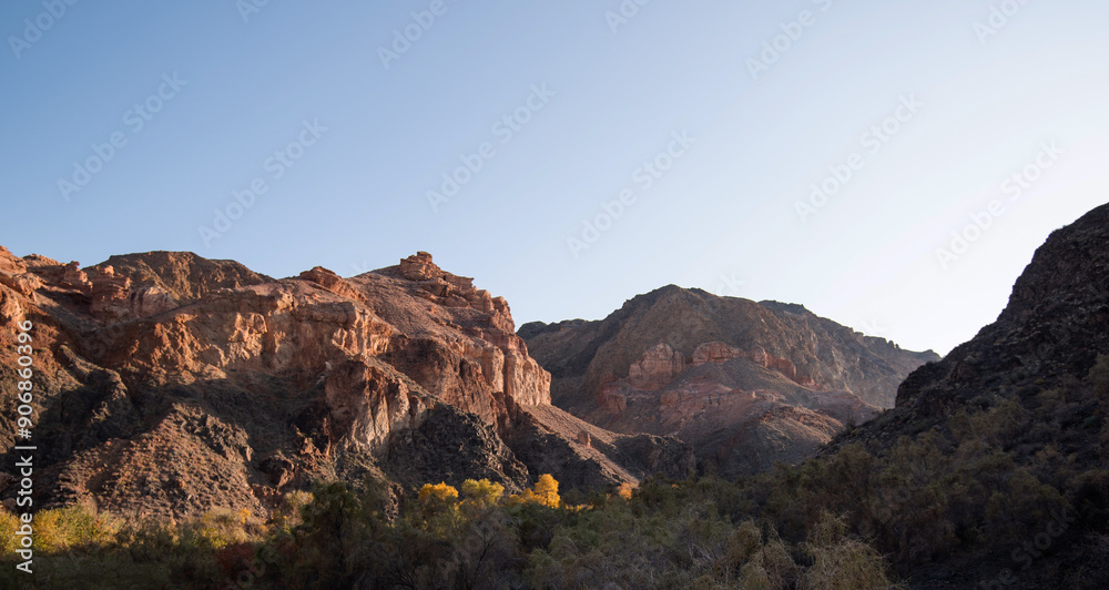 The morning sun illuminates a serene desert canyon with layered red rock cliffs and contrasting dark volcanic stones, interspersed with pockets of lush greenery The morning sun illuminates a serene desert canyon with layered red rock cliffs and contrasting dark volcanic stones, interspersed with pockets of lush greenery