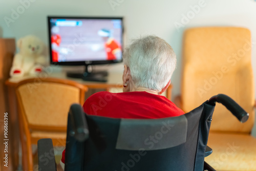 Senior woman with her back turned sitting in a wheelchair while enjoying watching TV in solitude at home
