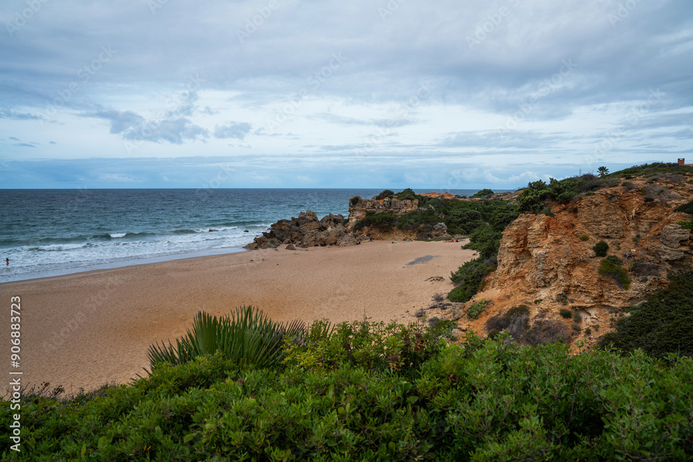 A secluded sandy beach bordered by rugged cliffs and Mediterranean shrubbery, with a solitary figure walking by the water under a partly cloudy sky.