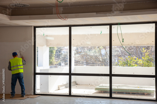 Construction worker smoothing drywall near a large window, showcasing expertise and attention to detail in a bright, professional setting.