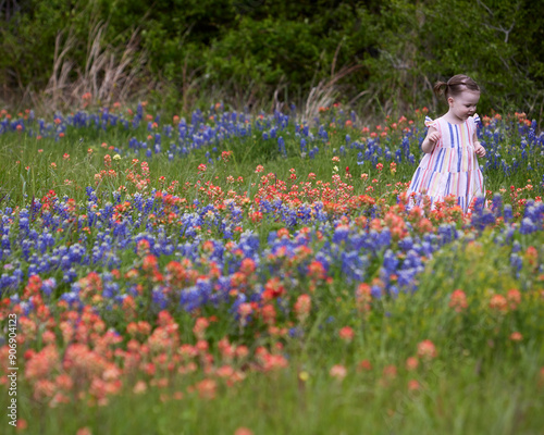 A young girl in a striped sundress smiles, looks down at the carpet of red, and blue wildflowers surrounding her.
