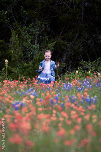 A little girl in a denim skirt and jacket a dark wood at her back, walks it a field of orange, red, and blue spring wildflowers.