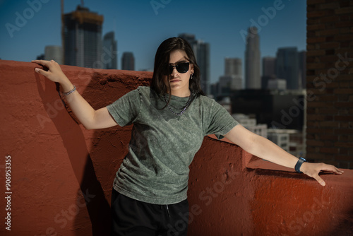 A person wearing a green shirt, black shorts, and sunglasses poses on a red rooftop wall with a cityscape of skyscrapers behind them, exuding confidence and a modern vibe. photo