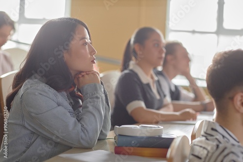 Side view of attentive young Asian female student attending university class sitting at desk in a row in lecture hall, copy space