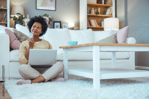 Thinking, laptop and woman with credit card on floor for subscription, payment or online shopping in living room. Decision, thoughtful and person for ideas, streaming service or investment in home photo