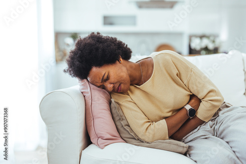 View of young woman suffering from stomachache on sofa at home. Woman sitting on bed and having stomach ache. Young woman suffering from abdominal pain while sitting on sofa at home photo