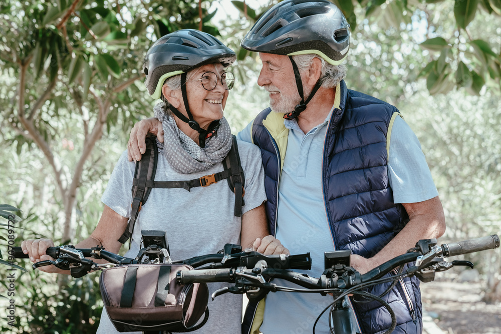 Fototapeta premium Active senior caucasian couple with electro bikes hug outdoors in the park. Two happy elderly people looking in the eyes enjoying nature and healthy lifestyle