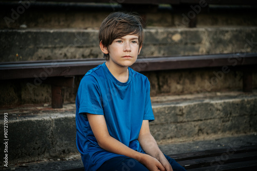 A young boy in a blue shirt gazes thoughtfully into the distance. The natural, blurred background features greenery, creating a serene outdoor setting.