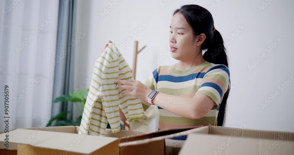 Handheld selective focus shot, Young Asian woman is sitting on the floor folding clothes into a cardboard box to prepare for a move. Moving house and lifestyle concept. Handheld selective focus shot, Young Asian woman is sitting on the floor folding clothes into a cardboard box to prepare for a move. Moving house and lifestyle concept.