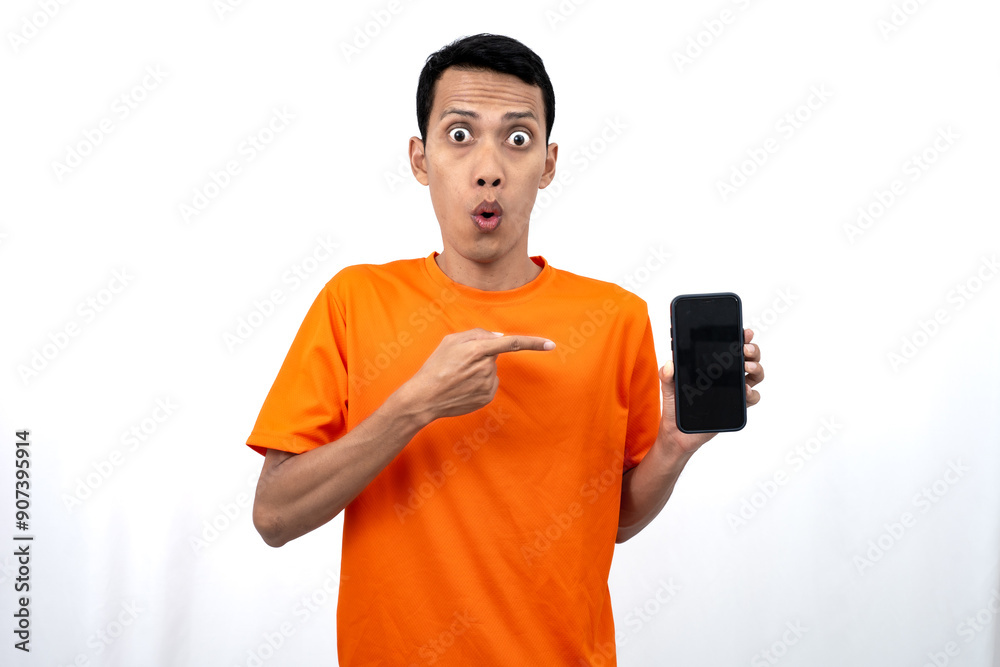 Portrait of a young Asian man wearing an orange t-shirt with a happy, surprised, pleased expression while pointing at a smartphone screen. Isolated on a white background. Portrait of a young Asian man wearing an orange t-shirt with a happy, surprised, pleased expression while pointing at a smartphone screen. Isolated on a white background.