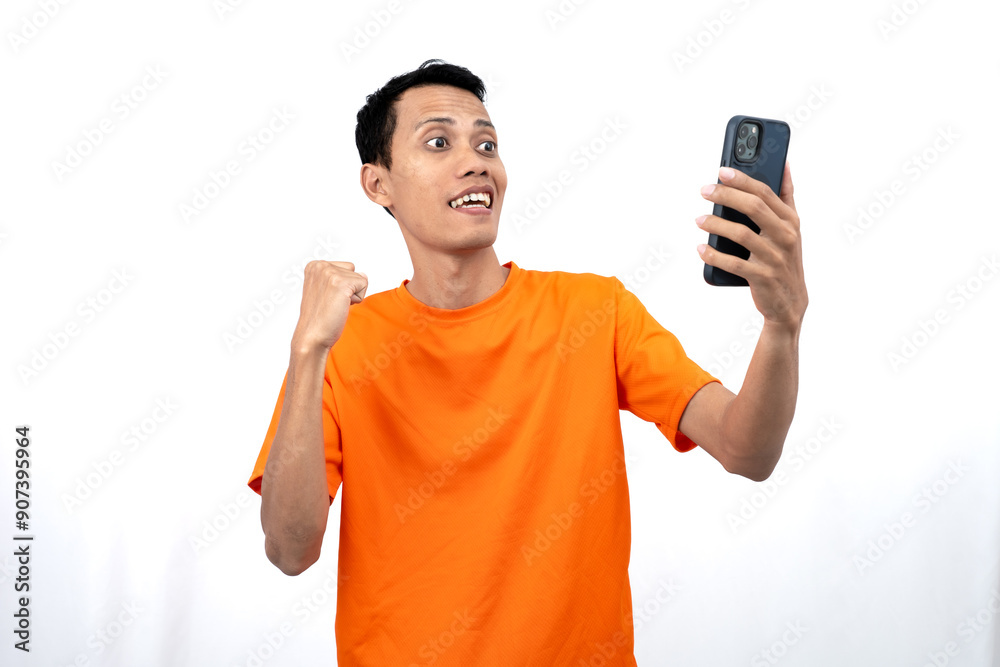 Portrait of a young Asian man wearing an orange t-shirt with a happy expression, surprised to get happy news via smartphone. Isolated on white background. Portrait of a young Asian man wearing an orange t-shirt with a happy expression, surprised to get happy news via smartphone. Isolated on white background.