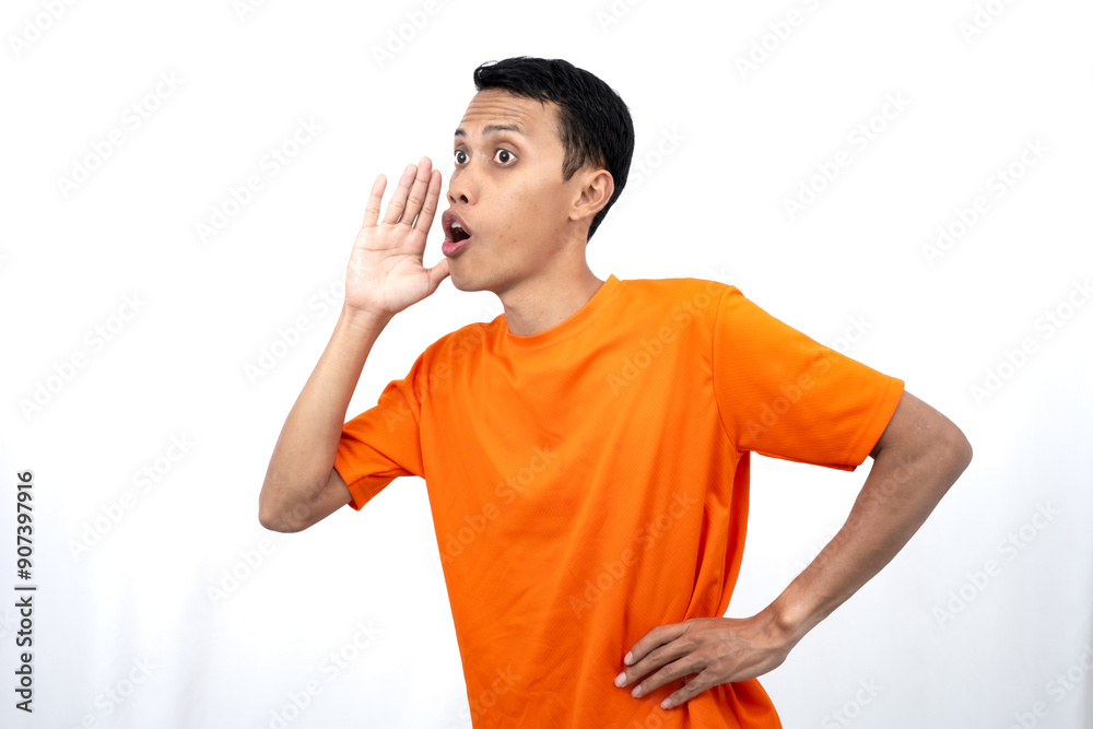 Portrait of a young Asian man wearing an orange t-shirt with his hand near his mouth whispering gossip and telling secrets while sharing news isolated on white background.