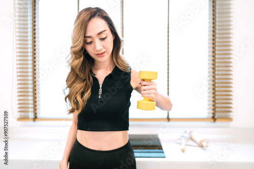 confident beautiful woman lifting exercise dumbbell in kitchen. gorgeous asian woman with home fitness concept holding dumbbell. beautiful motivated exerciser using light weight tool for arm muscle photo