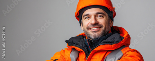 A man in an orange jacket and a hard hat is smiling. He is wearing a safety helmet and a jacket with reflective material