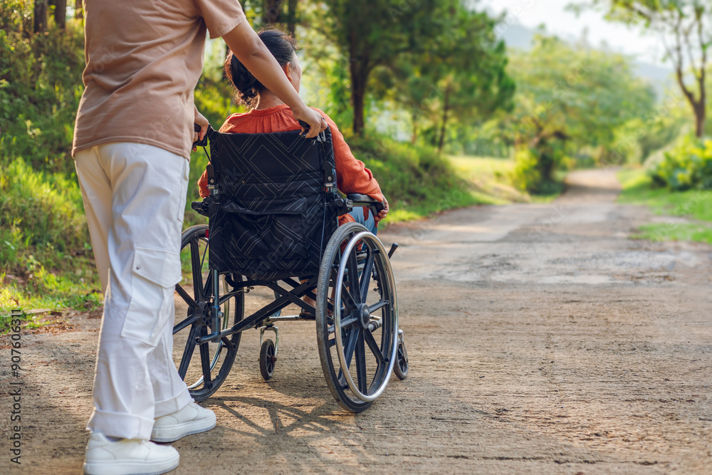 Woman sitting in wheelchair with his care helper walking on park trail outdoor morning sunrise mountain background. Rear view Woman sitting in wheelchair with his care helper walking on park trail outdoor morning sunrise mountain background. Rear view